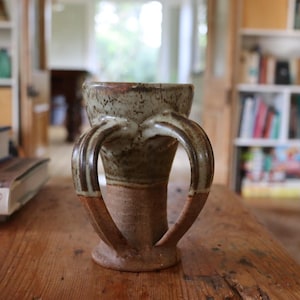 May include: A handmade ceramic vase with two curved handles. The vase has a speckled, light green and brown exterior with a reddish-brown base. It sits on a wooden table, with bookshelves and books in the background. The vase is approximately 18 cm tall.