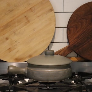 May include: A grey ceramic cooking pot with a lid and handle sits on a gas hob. Two wooden chopping boards, one light and one dark, are in the background. The boards are round with handles.