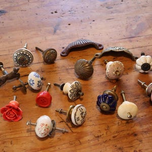 May include: A selection of decorative cabinet knobs and pulls in various styles and colours, including floral, geometric, and antique designs. The hardware is made of ceramic, metal, and other materials, displayed on a wooden surface.