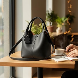 May include: A black leather handbag with a textured finish, featuring a top handle and a detachable shoulder strap. The bag has a small, decorative charm and is set on a wooden table near a window. A cup of coffee and a notebook are also on the table.