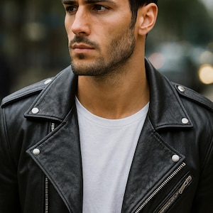May include: A black leather jacket with silver hardware, including a zipper and snap closures. The jacket is worn over a white crew neck t-shirt. The image is a close-up shot of the jacket and the person wearing it.