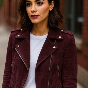 May include: A burgundy suede biker jacket with silver zippers and snap details. The jacket is worn over a white t-shirt. The model is wearing black jeans. The jacket has a classic asymmetrical design.