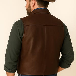 May include: A brown leather vest with a collared design. The vest is worn over a dark green long-sleeve shirt and blue jeans. A brown fedora hat completes the outfit.
