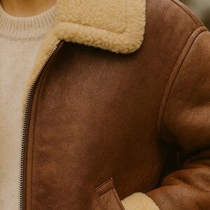 May include: A brown leather jacket with a cream-colored shearling collar and trim. The jacket has a zipper closure and a pocket on the front. The jacket is a classic style, suitable for cold weather.