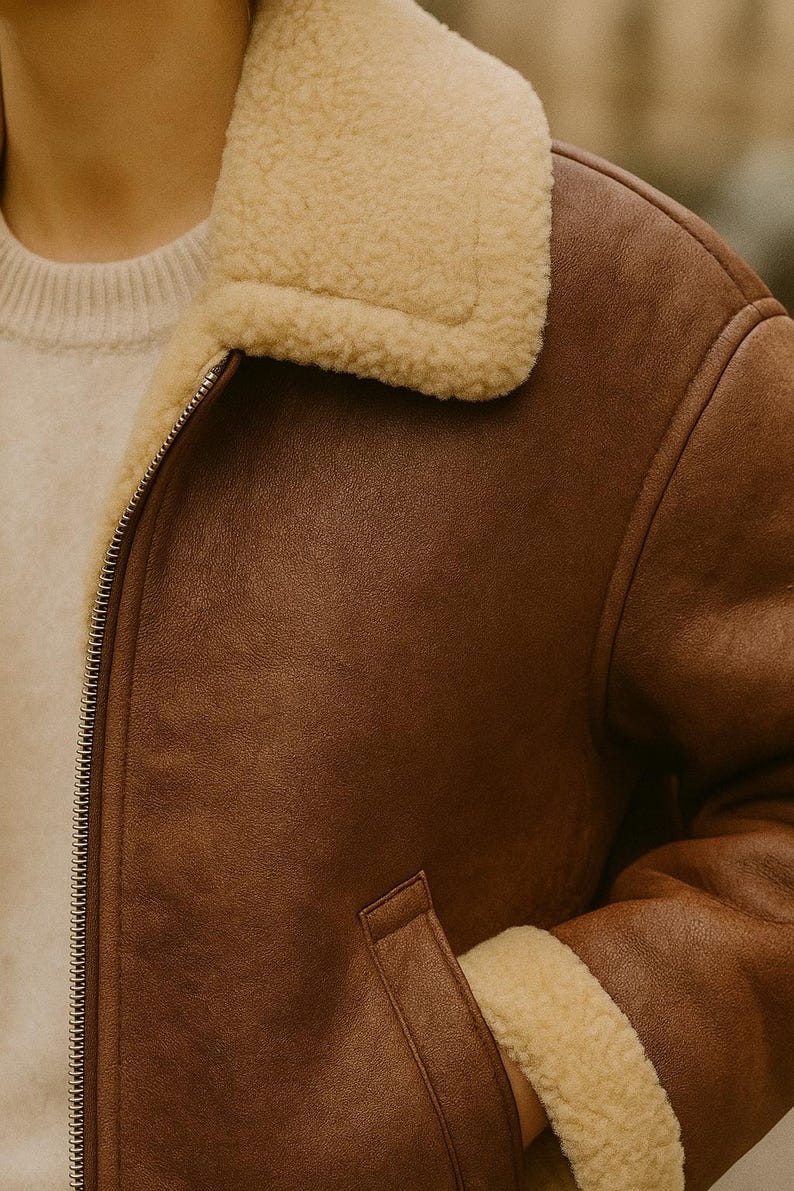 May include: A brown leather jacket with a cream-colored shearling collar and trim. The jacket has a zipper closure and a pocket on the front. The jacket is a classic style, suitable for cold weather.