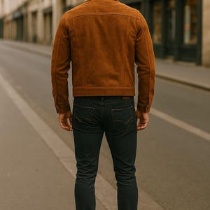 May include: A brown suede jacket and dark blue jeans. The jacket has a classic design with a collar and button closure. The jeans are a slim fit. The person is standing on a city street.