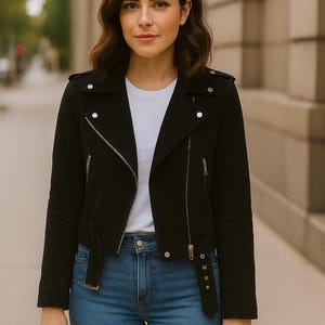 May include: A black jacket with silver zippers and snaps, paired with a white t-shirt and blue jeans. The jacket has a classic biker style with a belted waist. The outfit is completed with a black handbag.