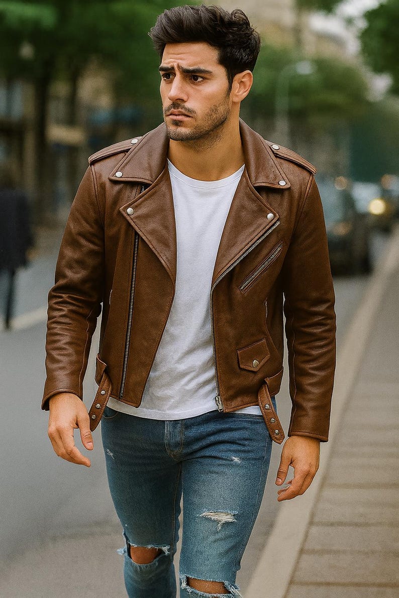 May include: A brown leather motorcycle jacket with a classic asymmetrical zipper design. The jacket features a notched collar, shoulder epaulets, and a small pocket. The person is wearing a white t-shirt and blue jeans.