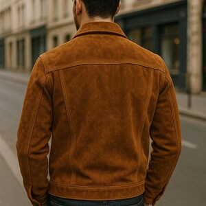 May include: A tan suede jacket with a classic collar and button closure. The jacket has a tailored fit and features visible stitching details. The person is wearing dark blue jeans. The image is taken outdoors.