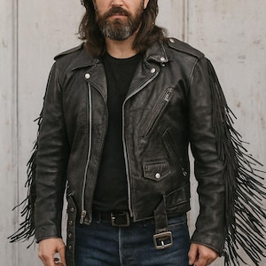 Puede incluir: Chaqueta de cuero negro con detalles de flecos. La chaqueta presenta un estilo motero clásico con cuello de solapa, bolsillos con cremallera y cinturón en la cintura. Las mangas tienen largos flecos. La persona lleva una camiseta negra y vaqueros azules.