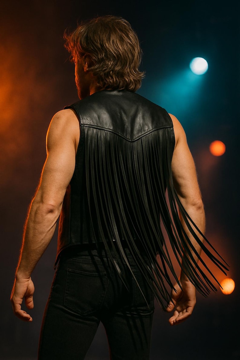 Op de afbeelding: Zwarte leren vest met lange franjes. Het vest heeft een klassieke V-hals en wordt gedragen met een zwarte jeans. De afbeelding is tegen een donkere achtergrond met oranje en blauwe spotlights, wat een dramatisch effect cre&euml;ert.