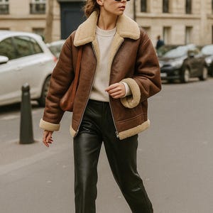 May include: A brown shearling jacket with a cream-colored lining and collar. The jacket is worn over a cream-colored sweater and paired with black leather pants. The outfit is completed with brown boots and sunglasses.