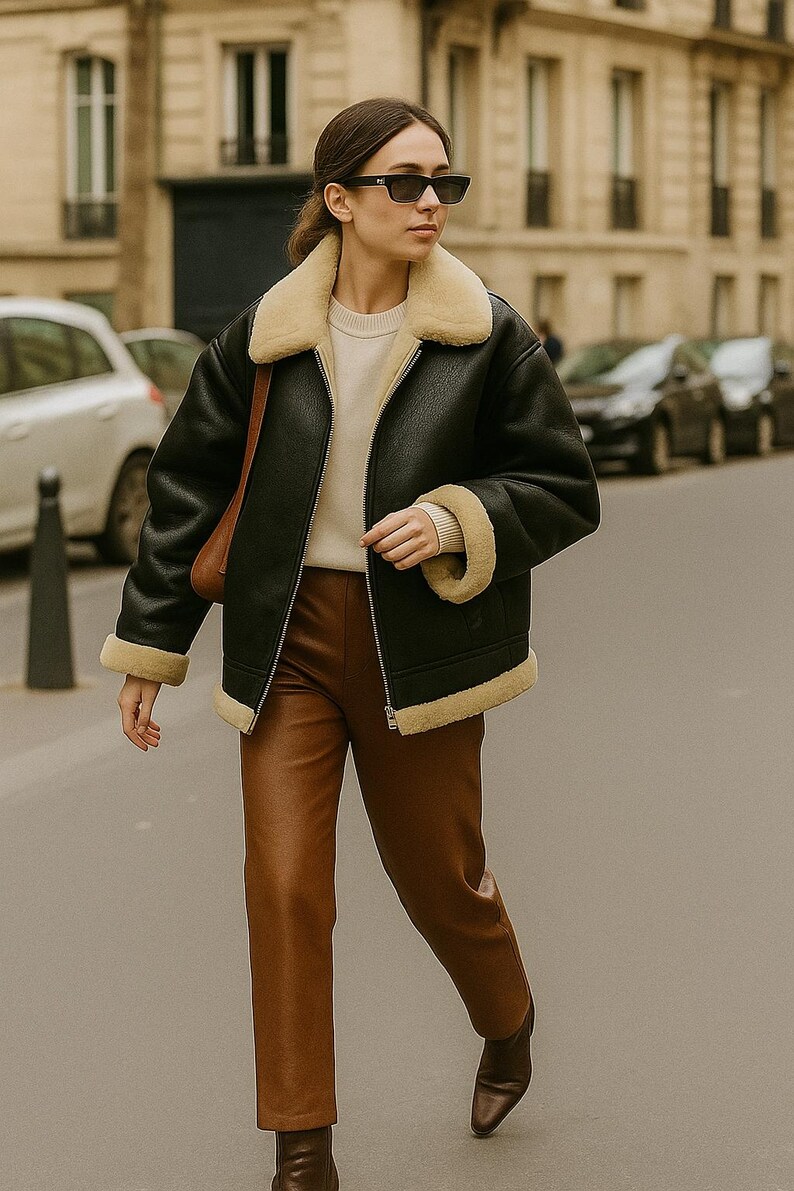 May include: A black shearling jacket with a cream-colored lining and trim, paired with brown leather pants. The jacket has a zipper closure and a cream-colored collar. The outfit is completed with brown leather boots.