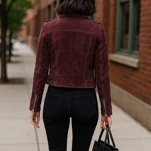 May include: A burgundy suede jacket with a zipper closure and black jeans. The jacket has a classic biker style with a tailored fit. A black leather handbag is carried in the right hand. The person is walking on a sidewalk.