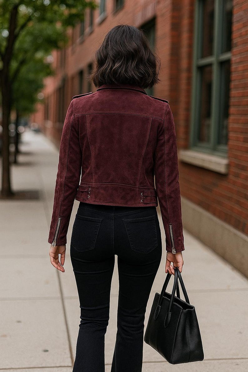 May include: A burgundy suede jacket with a zipper closure and black jeans. The jacket has a classic biker style with a tailored fit. A black leather handbag is carried in the right hand. The person is walking on a sidewalk.