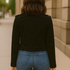 May include: A black jacket with a belt and blue jeans. The jacket has a classic design with a collar and shoulder accents. The jeans are a light blue wash. A black handbag is held in the hand.