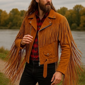May include: A tan suede jacket with long fringe detailing on the sleeves and chest. The jacket has a zipper closure, a small pocket, and a belt. The person is wearing a red and black plaid shirt and black jeans.