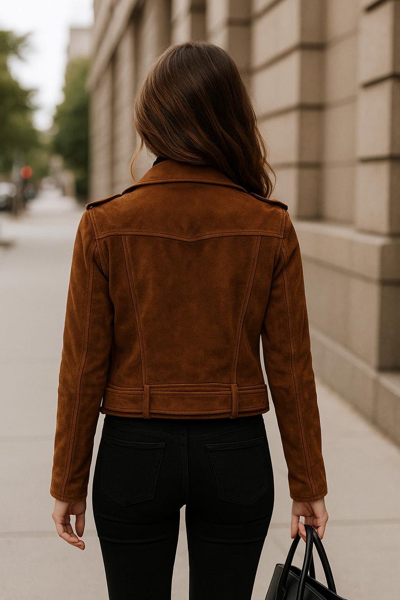 May include: A brown suede jacket with a classic design, featuring a notched collar, shoulder epaulets, and a belted waist. The jacket is paired with black jeans. The person is walking on a sidewalk.