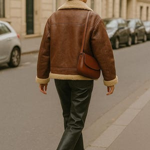 May include: A brown leather jacket with a shearling collar and cuffs, paired with black leather pants and a matching brown purse. The jacket has a classic aviator style. The person is walking on a city street.