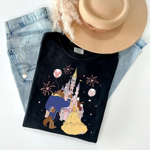 以下が含まれることがあります： お城の前で、花火と風船が描かれた、美女と野獣のカラフルなイラストが特徴の黒いTシャツ。明るいブルーのジーンズとタン色の帽子と組み合わせています。ゴールドのネックレスも確認できます。