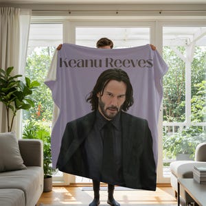 Könnte beinhalten: Eine hellviolette Fleecedecke mit einem großen Fotodruck von Keanu Reeves in einem schwarzen Anzug mit Krawatte. Der Name Keanu Reeves ist in dunkelbrauner Serifenschrift oben auf der Decke, mittig über dem Porträt, aufgedruckt.