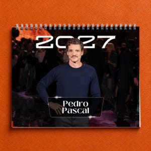 May include: A spiral-bound wall calendar for the year 2027 featuring a photograph of Pedro Pascal wearing a dark blue long-sleeve sweater. The text 2027 is printed in large white font at the top, and the name Pedro Pascal appears in a white box.