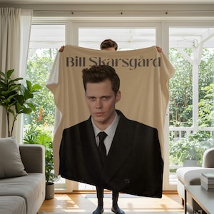 May include: A rectangular throw blanket featuring a large photographic print of Bill Skarsgård wearing a black suit, white shirt, and black tie. The name Bill Skarsgård is printed in dark gray serif font at the top against a light beige background.