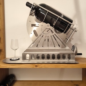 Könnte beinhalten: Ein Weinhalter aus Edelstahl mit einem einzigartigen mechanischen Design. Der Halter ist abgewinkelt und hält eine Flasche Rotwein. Ein Weinglas steht auf einem kleinen, runden Metalltablett. Der Halter befindet sich auf einem Holzregal.