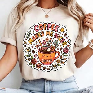 Pode incluir: Camiseta creme com um gráfico colorido. O gráfico inclui uma xícara de café sorrindo com a palavra "COFFEE" e a frase "MAKES ME NICER" em um design floral. O design também inclui flores, uma borboleta e grãos de café.