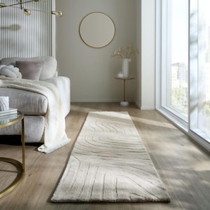 May include: A neutral-toned, rectangular area rug with a wave-like pattern. The rug is placed on a hardwood floor in a living room setting. The room also features a sofa, a side table, and a large window.