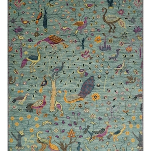 Puede incluir: Una alfombra turquesa con un patrón detallado de pájaros, árboles y flores. La alfombra presenta varias especies de aves en tonos naranja, morado, amarillo y azul, sobre un fondo de árboles estilizados y elementos florales. La alfombra tiene un borde con flecos.