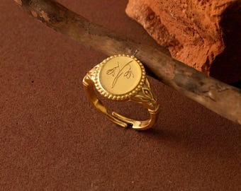 Puede incluir: Un anillo ajustable de color dorado con una cara redonda con un diseño floral. El anillo tiene un borde de cuentas y detalles ornamentados en la banda. El anillo se muestra sobre una superficie marrón con una rama de madera y un trozo de ladrillo.