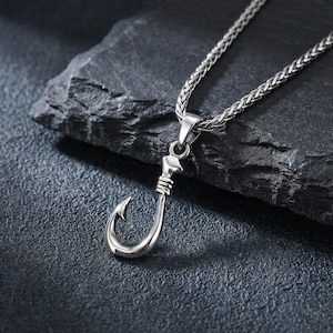 Könnte beinhalten: Eine silberfarbene Halskette mit einem Fischhaken-Anhänger. Der Haken ist mit einem strukturierten Design versehen und hängt an einer passenden Kette. Der Schmuck wird vor einem dunklen, strukturierten Hintergrund präsentiert, der den metallischen Glanz hervorhebt.