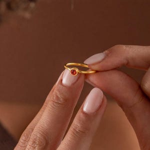 Op de afbeelding: Een gouden ring met een kleine, ronde, oranje edelsteen. De ring wordt tussen twee vingers gehouden, waarbij de eenvoudige band en de levendige kleur van de steen worden benadrukt. De ring is een sieraad.
