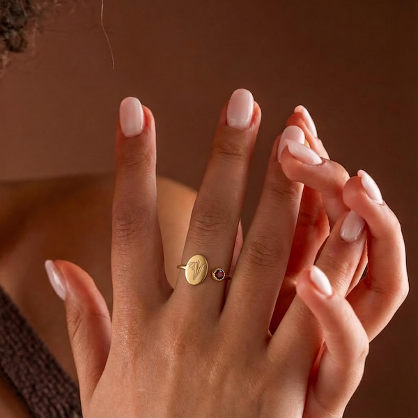 Pode incluir: Um anel de ouro com uma face oval gravada com a letra "V" e uma pequena pedra preciosa vermelha. O anel está em um dedo com esmalte rosa claro.