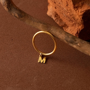 May include: A gold ring featuring a pendant with the letter "M". The ring is displayed on a brown surface, with a wooden branch and a piece of reddish-brown stone in the background.