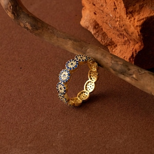 May include: A gold-colored ring with a repeating pattern of blue and white circular designs. The ring is set against a brown background with a wooden branch and a piece of red brick.