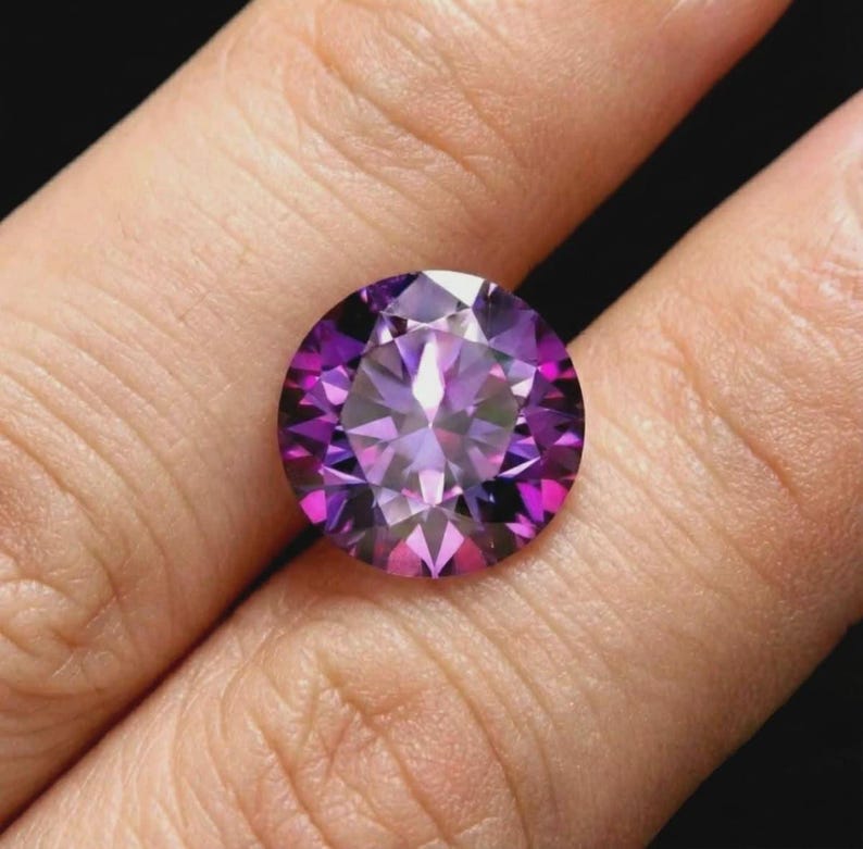 K&ouml;nnte beinhalten: Ein runder, facettierter Edelstein mit Brillantschliff. Der Edelstein zeigt eine Reihe von lilafarbenen Farbt&ouml;nen, von tiefem Violett bis zu hellem Lavendel, mit einer hellen, reflektierenden Oberfl&auml;che. Der Edelstein wird zwischen Fingern vor dunklem Hintergrund gehalten.