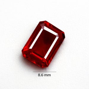 May include: A faceted, rectangular, deep red gemstone. The gem is 8.6 mm long. The cut of the stone creates multiple reflective surfaces. The gem is set against a white background.