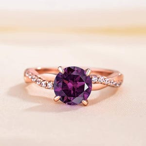 K&ouml;nnte beinhalten: Ros&eacute;goldener Ring mit einem gro&szlig;en, runden, tiefvioletten Edelstein. Das Band hat ein verdrehtes Design mit kleinen, klaren Akzentsteinen. Der Ring ist vor einem weichen, neutralen Hintergrund platziert, der die lebendige Farbe des zentralen Steins hervorhebt.