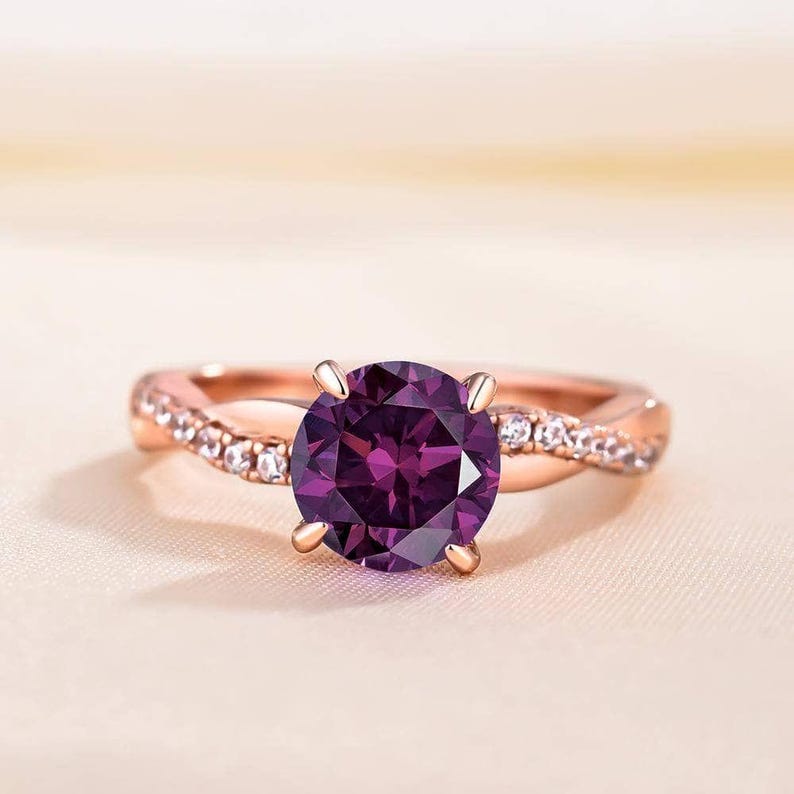 K&ouml;nnte beinhalten: Ros&eacute;goldener Ring mit einem gro&szlig;en, runden, tiefvioletten Edelstein. Das Band hat ein verdrehtes Design mit kleinen, klaren Akzentsteinen. Der Ring ist vor einem weichen, neutralen Hintergrund platziert, der die lebendige Farbe des zentralen Steins hervorhebt.