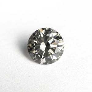 Könnte beinhalten: Ein runder Edelstein im Brillantschliff mit dunkelgrauem, metallischem Aussehen. Der Stein weist zahlreiche dreieckige und rautenförmige Facetten auf, die ein komplexes Muster aus Licht und Schatten erzeugen und eine hohe Klarheit widerspiegeln.