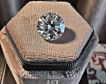 May include: A round, brilliant-cut clear gemstone with high light refraction, resting on a hexagonal, beige velvet display box. The text 8mm and 2 CT is printed on the lower portion of the velvet surface, indicating the size and weight.