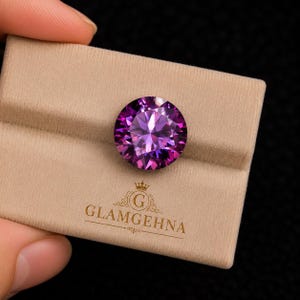 K&ouml;nnte beinhalten: Ein runder, facettierter violetter Edelstein, m&ouml;glicherweise Amethyst, auf einer beigen Samtoberfl&auml;che pr&auml;sentiert. Der Edelstein zeigt seinen aufwendigen Schliff und seine Farbe. Das Logo "GLAMGEHNA" ist in Gold auf der Oberfl&auml;che gedruckt.