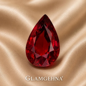 May include: A faceted, pear-shaped, deep red gemstone. The gem is set against a backdrop of a golden, satin-like fabric. The word "GLAMGEHNA" is printed in white at the bottom of the image.