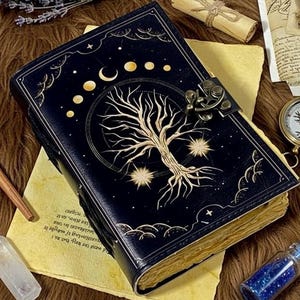 Puede incluir: Un diario de cuero azul oscuro con un cierre de latón. La portada presenta un diseño de árbol dorado dentro de un círculo, con fases lunares y estrellas. El diario está abierto, revelando papel envejecido.