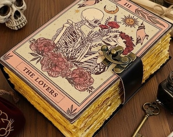 May include: A vintage-style journal with a black leather strap and clasp. The cover features a skeleton couple illustration with floral accents and the text "THE LOVERS". Other items include a skull, key, inkwell, and feather.
