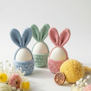 May include: Three pastel-colored crochet Easter egg holders shaped like bunnies. The holders are blue, green, and pink, each with a white egg inside. A yellow crochet ball and a wooden tree of life button are also present, along with flowers.