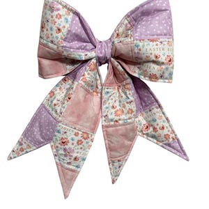 May include: A large, quilted bow in shades of pink and lavender, featuring floral and polka dot patterns. The bow has a patchwork design with the words "Easter Blessings" and "Rejoice" printed on it. The bow is tied in the center.