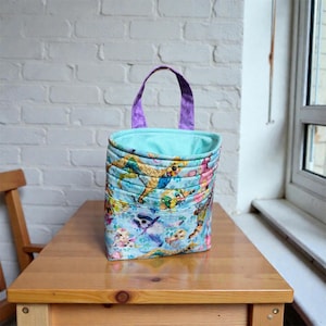 May include: A quilted fabric tote bag with a light blue background and a colorful dog print. The bag has a purple handle and a light blue interior. The tote is sitting on a wooden table.